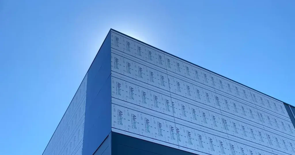 Modern building under construction with blue exterior panels, large windows, and scaffolding along the corner against a clear blue sky, including branded construction wrap.